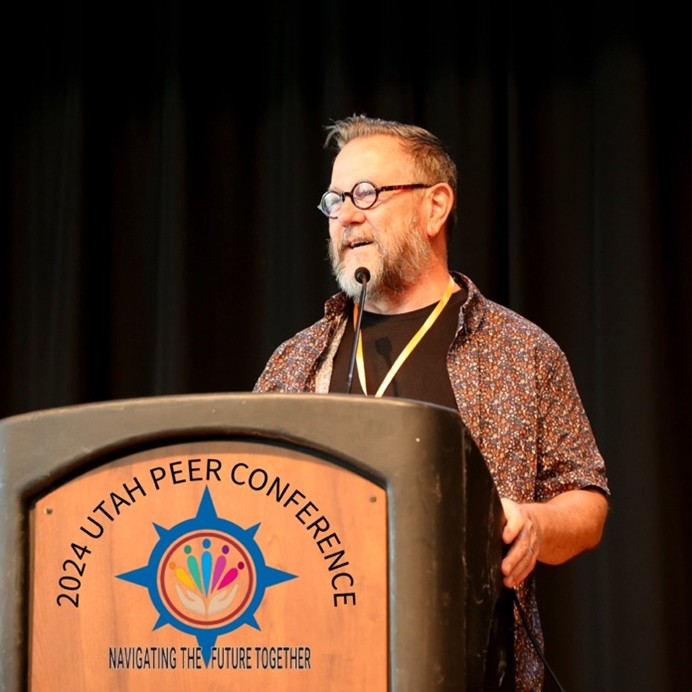 Donald McDonald standing behind a podium that has a logo which says, "2020 Utah Peer conference. Navigating the Future Together."