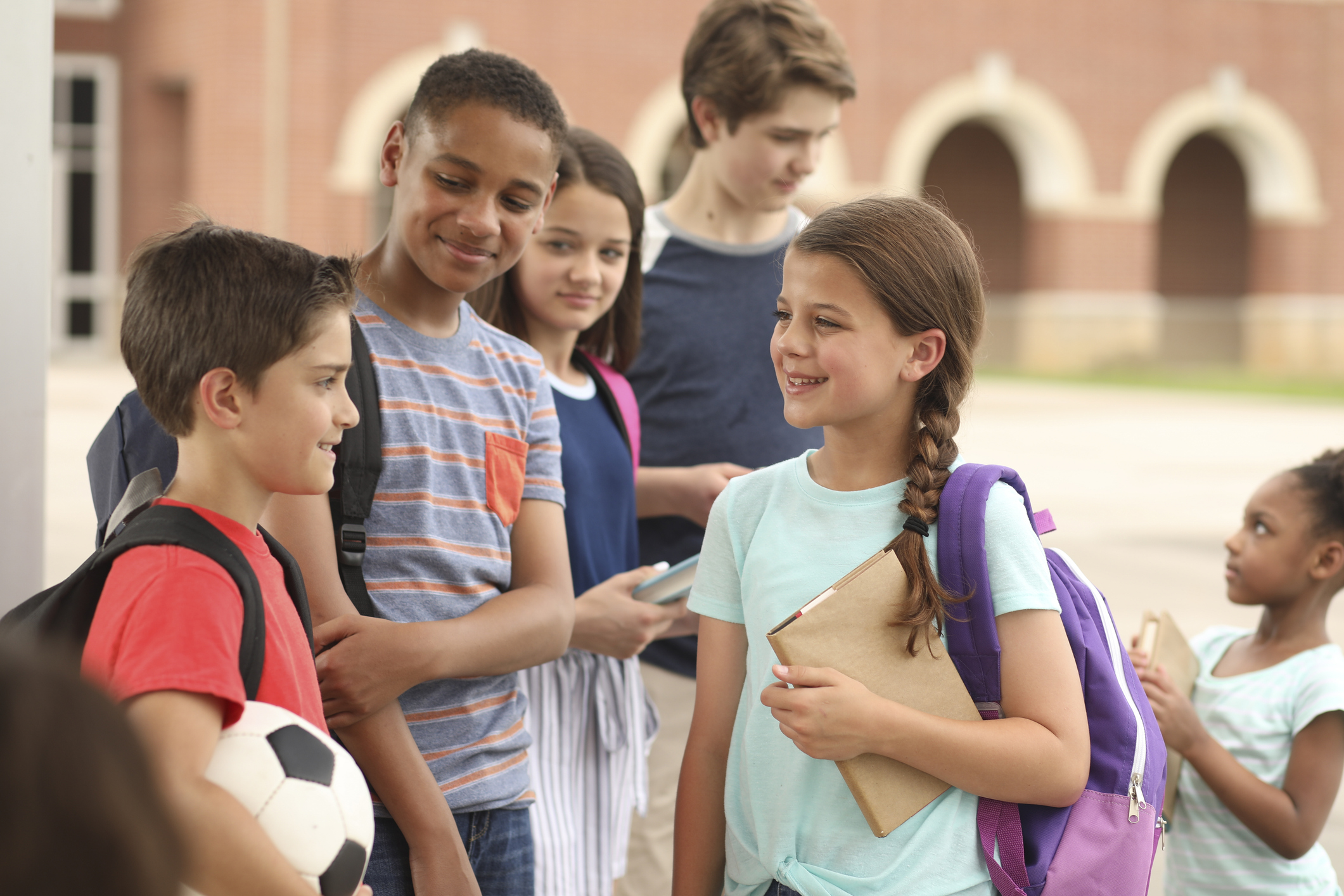 Group of six children and youths of different ages standing outside with backpacks and talking to each other