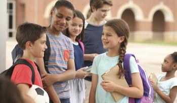 Group of six children and youths of different ages standing outside with backpacks and talking to each other